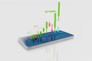A white smartphone lies on a light grey surface, displaying a 3D candlestick chart with green and red bars rising from its screen. Above the chart, the text 'BUY SIGNAL' is prominently displayed in green, indicating a trading opportunity.
