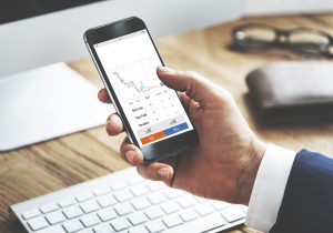 A close-up of a hand holding a smartphone displaying a Forex trading application. The phone screen shows a line graph with buy and sell options, and in the blurred background, a computer keyboard and a wallet with glasses are visible on a wooden desk.