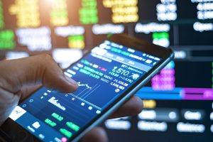 A close-up of a hand holding a smartphone displaying a trading application. The screen shows a financial chart with real-time data, including a balance of 1,510.78, against a blurred background of illuminated digital displays with more financial information.
